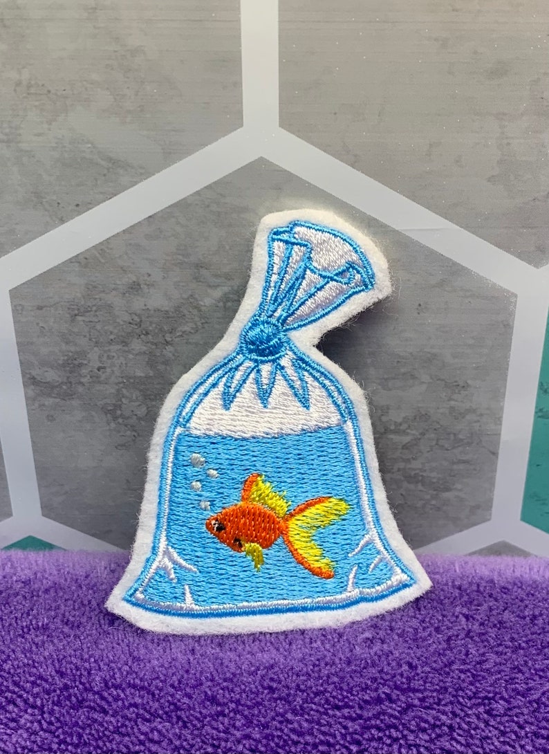 May include: An embroidered patch featuring a goldfish inside a clear plastic bag filled with water. The fish is orange with yellow fins, and the bag is tied at the top with white thread. The design includes small white bubbles in the blue water.