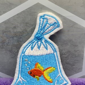 May include: An embroidered patch featuring a goldfish swimming inside a clear plastic bag filled with water. The design includes blue water, a bright orange and yellow fish, small white bubbles, and a tied top section with white highlights.