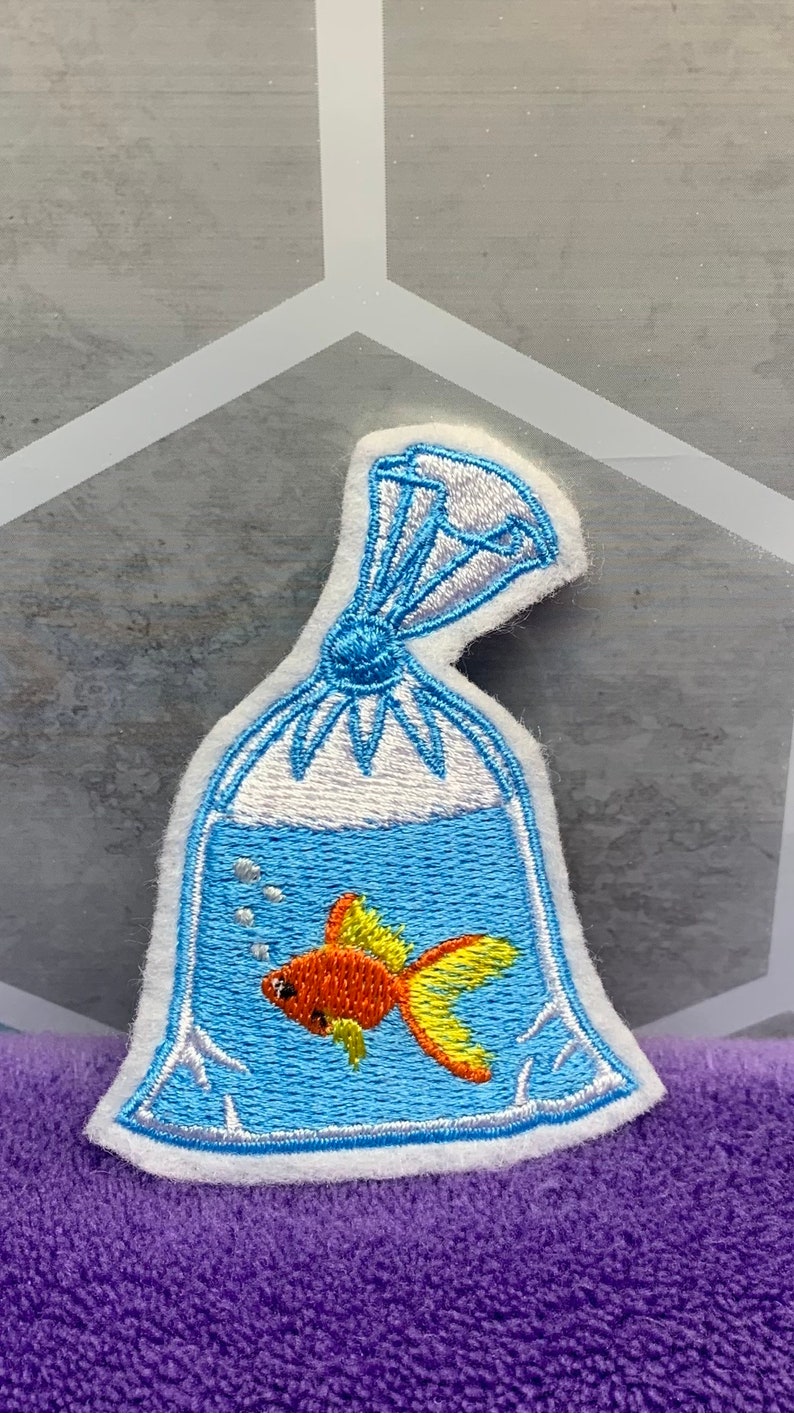 May include: An embroidered patch featuring a goldfish swimming inside a clear plastic bag filled with water. The design includes blue water, a bright orange and yellow fish, small white bubbles, and a tied top section with white highlights.
