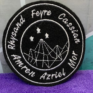May include: Black and white embroidered patch with a circle design. The circle features a mountain range with three stars above it. The words "Rhysand", "Feyre", "Cassian", "Amren", "Azriel", and "Mor" are written around the circle.