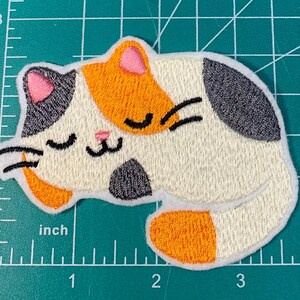 Op de afbeelding: Een geborduurde patch van een slapende kat, met witte, oranje en zwarte kleuren. De kat heeft zijn ogen dicht en een kleine roze neus.