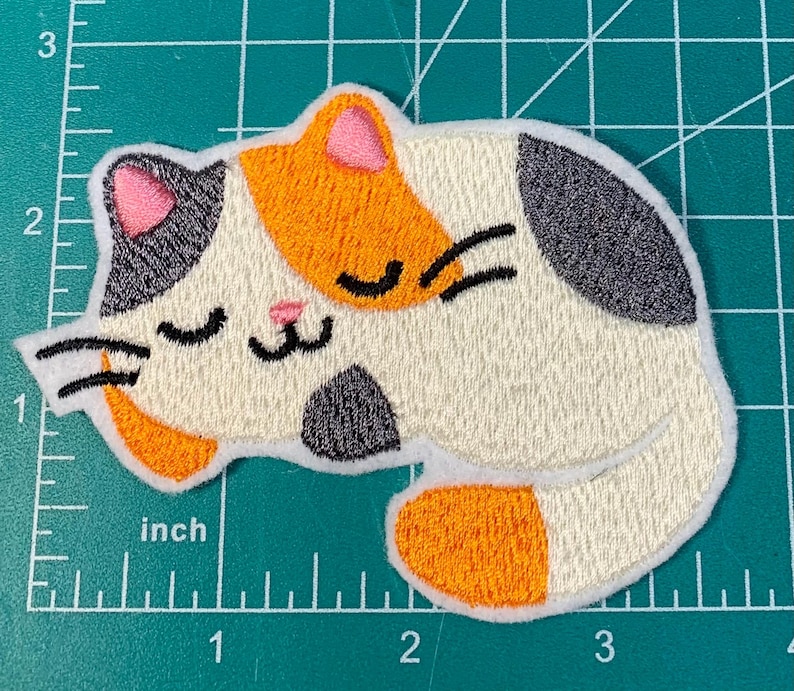 Op de afbeelding: Een geborduurde patch van een slapende kat, met witte, oranje en zwarte kleuren. De kat heeft zijn ogen dicht en een kleine roze neus.