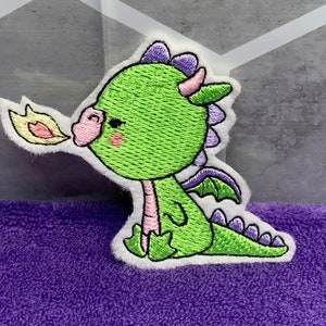 May include: An embroidered patch of a cartoon dragon, green with purple horns and wings, blowing a yellow flame from its mouth.
