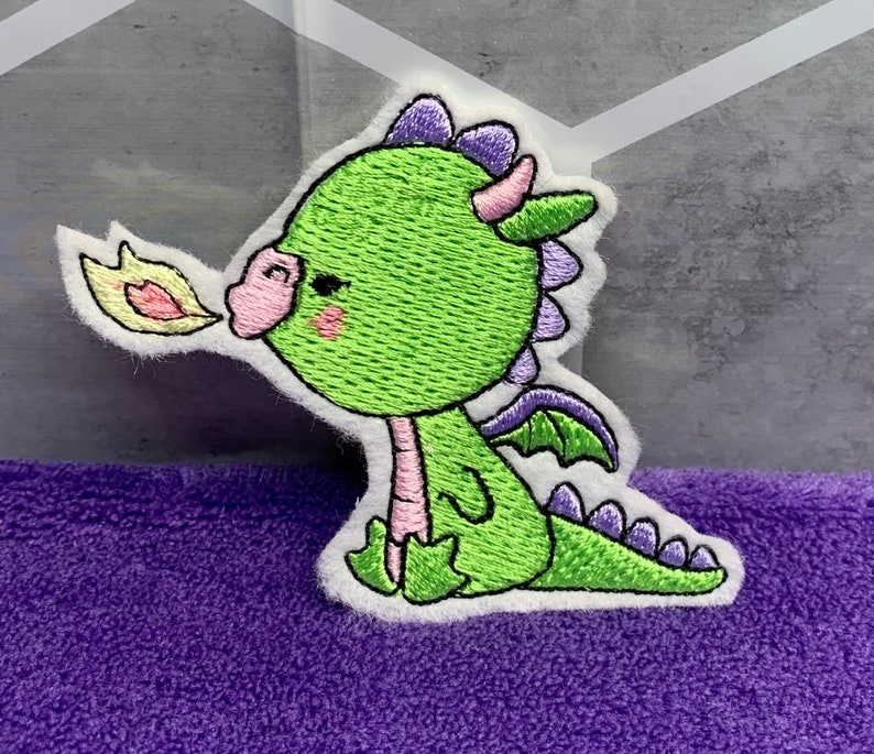 May include: An embroidered patch of a cartoon dragon, green with purple horns and wings, blowing a yellow flame from its mouth.