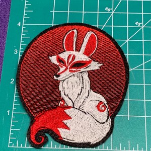 May include: Embroidered patch featuring a white fox with red accents, set against a red circular background. The fox has a detailed face and a curled tail. The patch is approximately 3 inches in height.