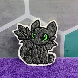May include: A black and green embroidered patch of a cartoon dragon with large eyes. The dragon is sitting on its haunches with its wings folded behind it.