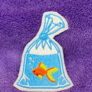 May include: An embroidered patch featuring a clear plastic bag filled with water and a small orange goldfish with yellow fins. The bag is tied at the top with blue thread and includes white embroidered bubbles floating above the fish.