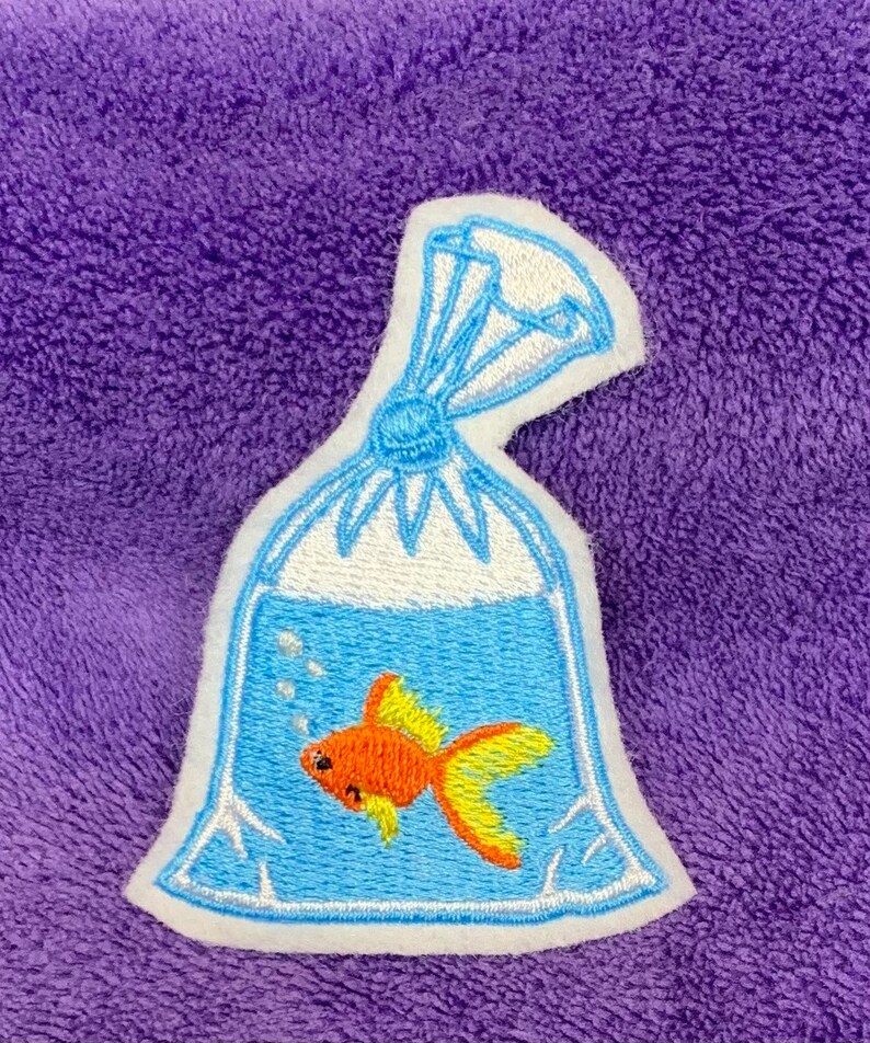 May include: An embroidered patch featuring a clear plastic bag filled with water and a small orange goldfish with yellow fins. The bag is tied at the top with blue thread and includes white embroidered bubbles floating above the fish.