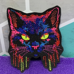 May include: Embroidered black cat patch with neon pink, blue, and green details. The cat's eyes are bright yellow and the patch has a dripping effect.