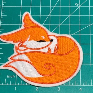 May include: Embroidered patch featuring a sleeping fox in shades of orange, white, and black. The fox is curled up, with its tail wrapped around its body. The patch is approximately 3 inches wide.