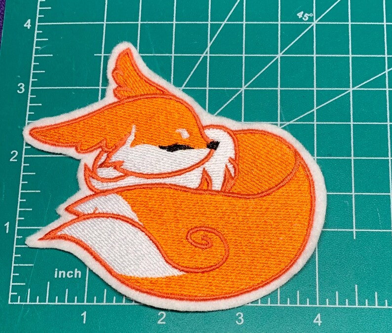 May include: Embroidered patch featuring a sleeping fox in shades of orange, white, and black. The fox is curled up, with its tail wrapped around its body. The patch is approximately 3 inches wide.