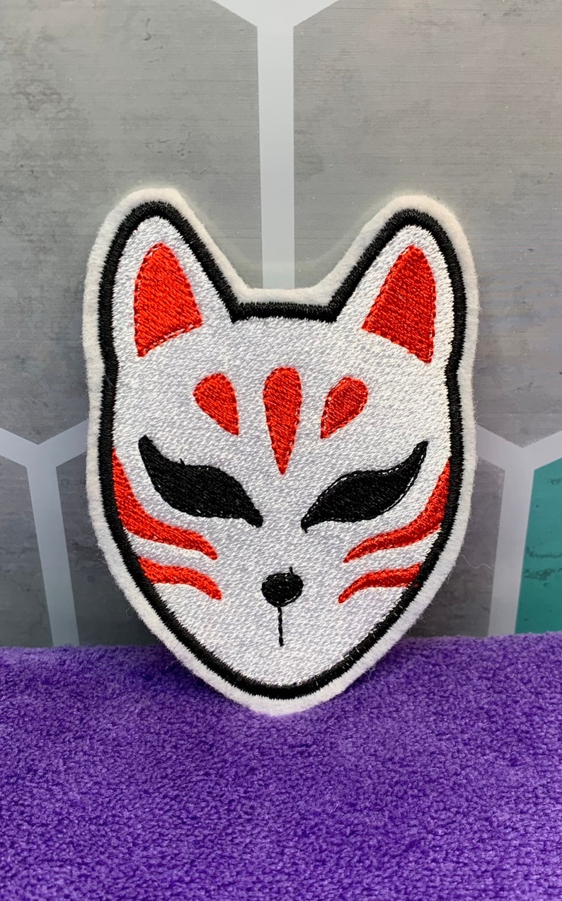May include: White embroidered patch with a red and black fox mask design. The mask has a stylized fox face with red ears, eyes, and markings.