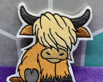 May include: A brown and white embroidered patch of a cartoon Highland cow with a fluffy mane. The cow is sitting with its legs tucked under its body.