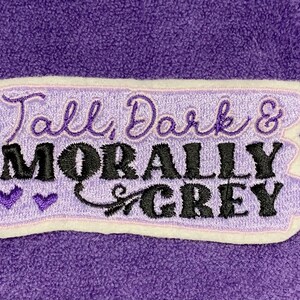 May include: A lavender embroidered patch with the words "Tall, Dark & Morally Grey" in black and lavender script. The patch has a white border and two purple heart accents, set against a purple fabric background.