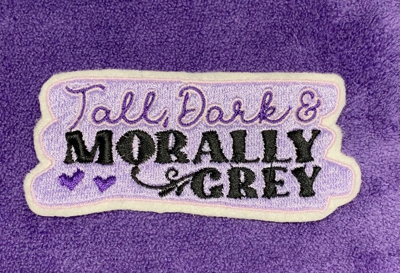 May include: A lavender embroidered patch with the words "Tall, Dark & Morally Grey" in black and lavender script. The patch has a white border and two purple heart accents, set against a purple fabric background.