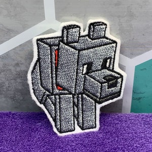 May include: Embroidered patch featuring a pixelated wolf design in shades of gray, black, and white. The wolf has a red accent on its chest and is outlined with white stitching. The patch is on a purple surface.