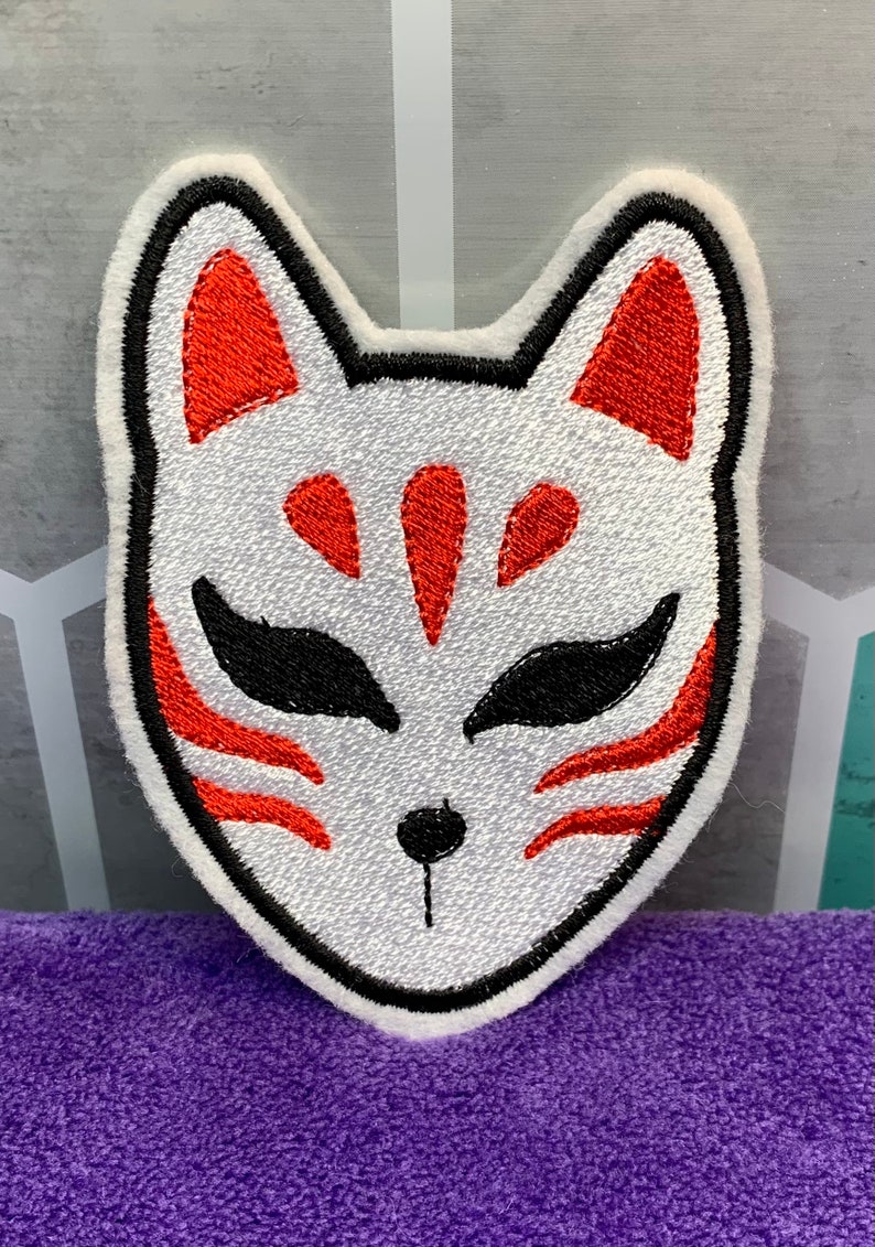 May include: White embroidered patch with a red and black fox mask design. The mask has a stylized fox face with red markings and black eyes.