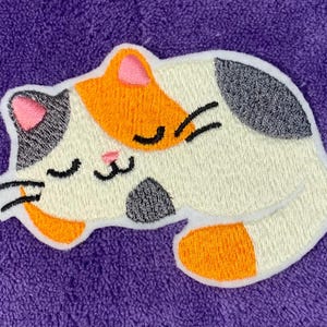 Op de afbeelding: Een geborduurde patch van een slapende kat, met witte, oranje en grijze kleuren. De kat heeft zijn ogen dicht en een roze neus.