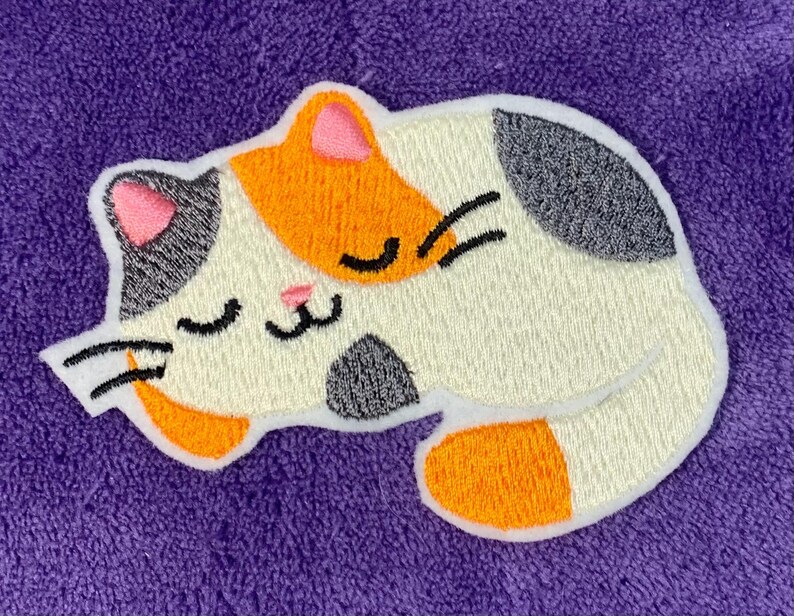 Op de afbeelding: Een geborduurde patch van een slapende kat, met witte, oranje en grijze kleuren. De kat heeft zijn ogen dicht en een roze neus.
