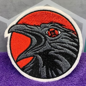 May include: Embroidered patch featuring a raven's head with an open beak, set against a red background within a white-bordered circle. The raven is detailed in shades of gray and black, with a red eye. Ideal for jackets, bags, or other fabric items.