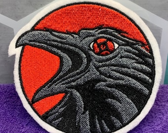 May include: Embroidered patch featuring a raven's head with an open beak, set against a red background within a white-bordered circle. The raven is detailed in shades of gray and black, with a red eye. Ideal for jackets, bags, or other fabric items.