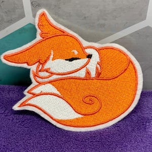 May include: Embroidered patch featuring a sleeping fox in shades of orange, white, and black. The fox is curled up, with its tail wrapped around its body. The patch has a white border and is likely for clothing or accessories.