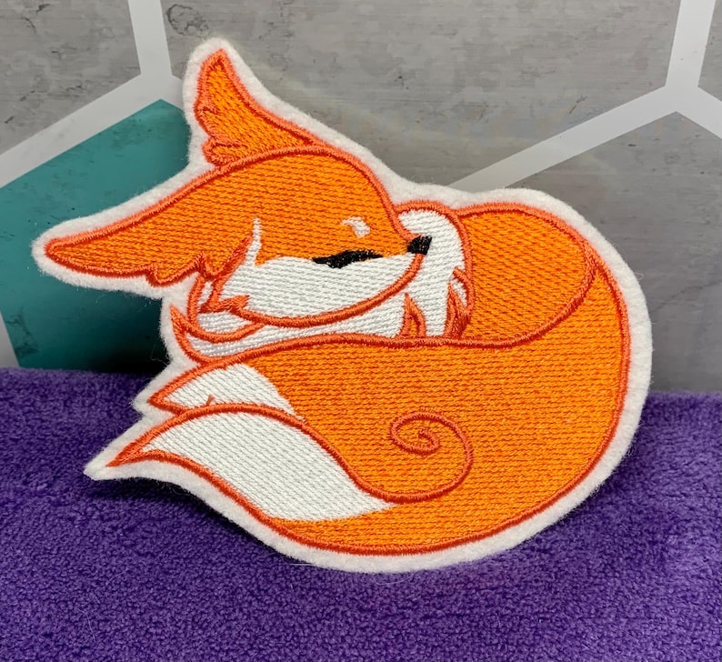 May include: Embroidered patch featuring a sleeping fox in shades of orange, white, and black. The fox is curled up, with its tail wrapped around its body. The patch has a white border and is likely for clothing or accessories.