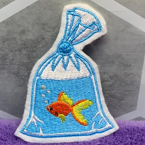May include: An embroidered patch featuring a clear plastic bag filled with blue water and a small orange goldfish with yellow fins. The bag is tied at the top with blue thread and includes white embroidered bubbles inside the water section.
