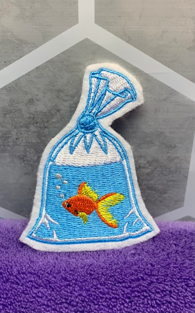 May include: An embroidered patch featuring a clear plastic bag filled with blue water and a small orange goldfish with yellow fins. The bag is tied at the top with blue thread and includes white embroidered bubbles inside the water section.