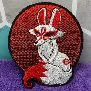 May include: Embroidered patch featuring a white fox with red accents, set against a red circular background. The fox has a mask-like design and a curled tail. The patch is outlined in black.