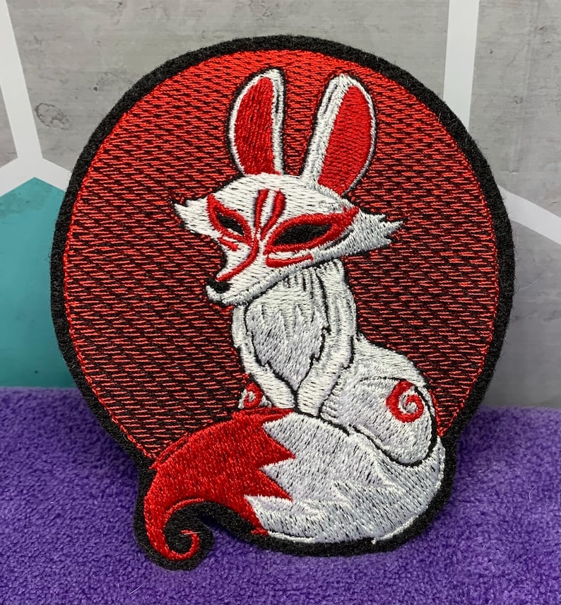 May include: Embroidered patch featuring a white fox with red accents, set against a red circular background. The fox has a mask-like design and a curled tail. The patch is outlined in black.