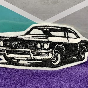 May include: Embroidered patch featuring a classic black and white car design. The patch has a white border and depicts a vintage coupe with detailed headlights, wheels, and body lines. The patch is on a purple fabric.