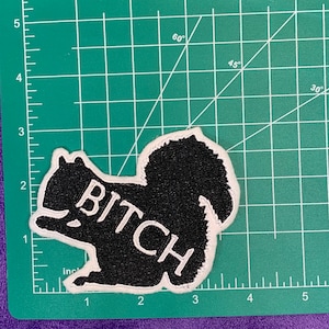 May include: Black and white embroidered patch of a squirrel with the word "BITCH" written across its body.