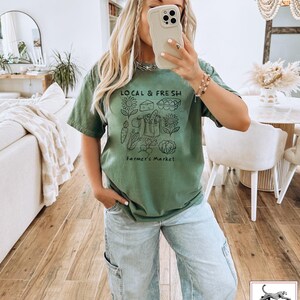 May include: Olive green t-shirt with a black graphic that reads "LOCAL & FRESH" above a farmer's market illustration. The illustration includes vegetables, cheese, eggs, and sunflowers. The text "Farmer's Market" is below the illustration.