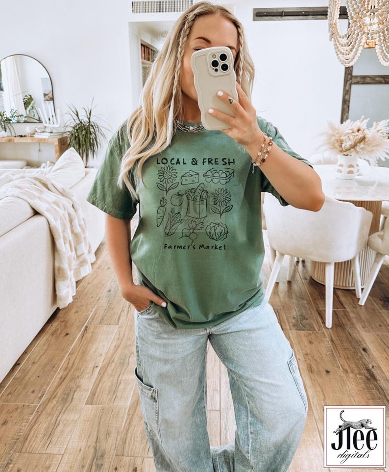 May include: Olive green t-shirt with a black graphic that reads "LOCAL & FRESH" above a farmer's market illustration. The illustration includes vegetables, cheese, eggs, and sunflowers. The text "Farmer's Market" is below the illustration.