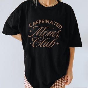 Op de afbeelding: Zwart oversized T-shirt met de tekst "caffeinated moms club" in bruine cursieve letters met sterren aan beide kanten van de tekst.