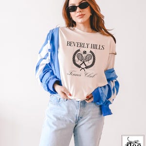 Puede incluir: Una camiseta blanca con un dise&ntilde;o gr&aacute;fico negro que dice "Beverly Hills Tennis Club" con un dise&ntilde;o de raqueta de tenis y pelota.