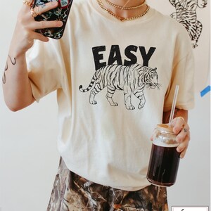 May include: A cream colored t-shirt with a black tiger graphic and the word "EASY" printed on the front. The model is wearing the shirt with camouflage shorts.