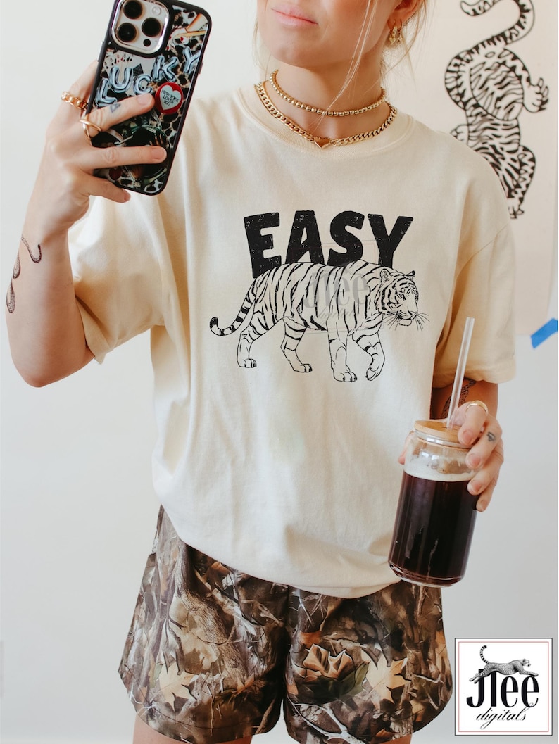 May include: A cream colored t-shirt with a black tiger graphic and the word "EASY" printed on the front. The model is wearing the shirt with camouflage shorts.
