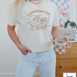 Op de afbeelding: Een wit T-shirt met de tekst "CAFFEINATED Moms Club" in bruine letters. Het shirt wordt gedragen door een persoon met lichtblauwe jeans.
