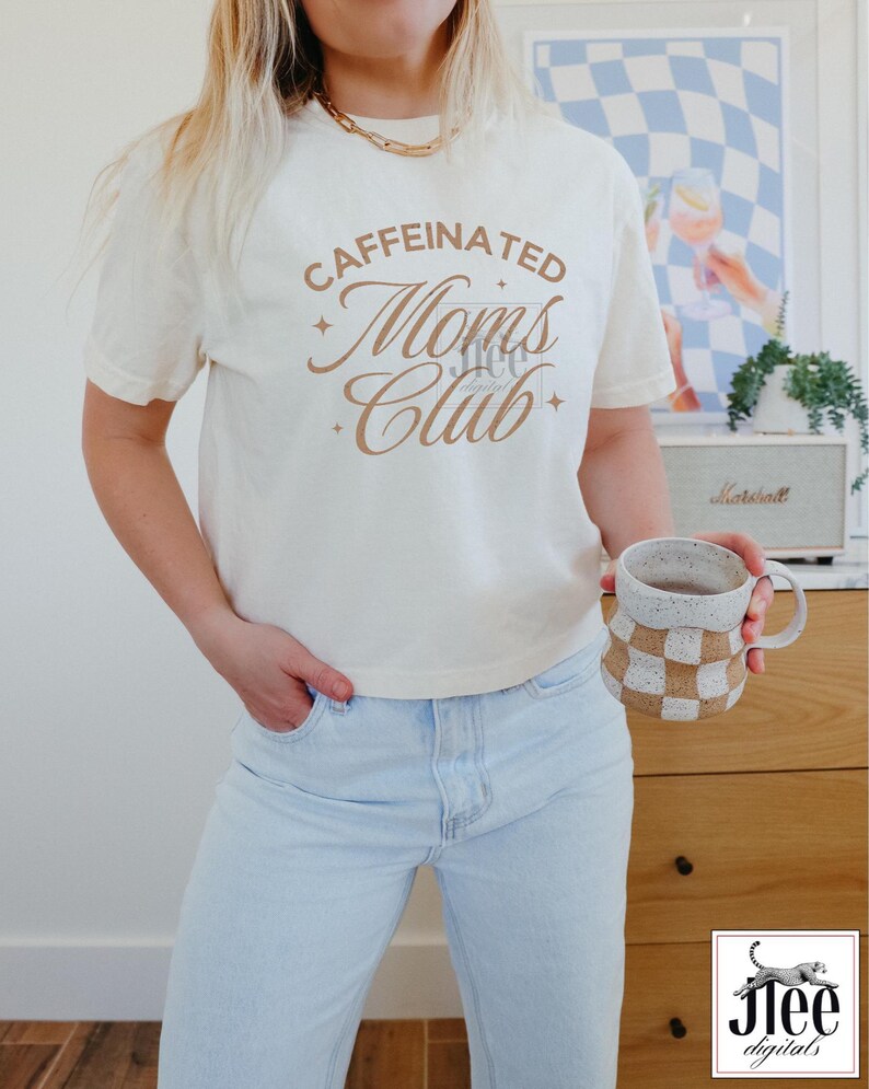 Op de afbeelding: Een wit T-shirt met de tekst "CAFFEINATED Moms Club" in bruine letters. Het shirt wordt gedragen door een persoon met lichtblauwe jeans.