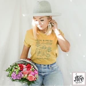 May include: Mustard yellow t-shirt with the text "LOCAL & FRESH" and a farmer's market graphic. The person is wearing a gray hat and blue jeans, holding a bouquet of flowers with red, pink, and purple blooms.