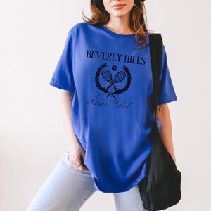 Puede incluir: Una camiseta azul real con un dise&ntilde;o gr&aacute;fico negro que dice "Beverly Hills Tennis Club" con un logotipo de raqueta de tenis y pelota.
