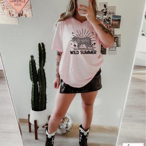 May include: Pink t-shirt with a black and white leopard print graphic and the text "Wild Summer". The model is wearing a black skirt, cowboy boots, and a white baseball cap with the text "Cowgirl Cowboy" on it.