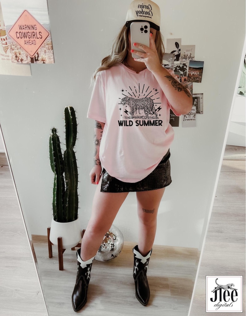 May include: Pink t-shirt with a black and white leopard print graphic and the text "Wild Summer". The model is wearing a black skirt, cowboy boots, and a white baseball cap with the text "Cowgirl Cowboy" on it.