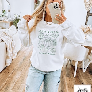 May include: White sweatshirt with the text "LOCAL & FRESH" and a green illustration of farmer's market produce. The design includes sunflowers, cheese, eggs, carrots, apples, a basket of vegetables, and a cabbage. The words "Farmer's Market" are below the illustration.