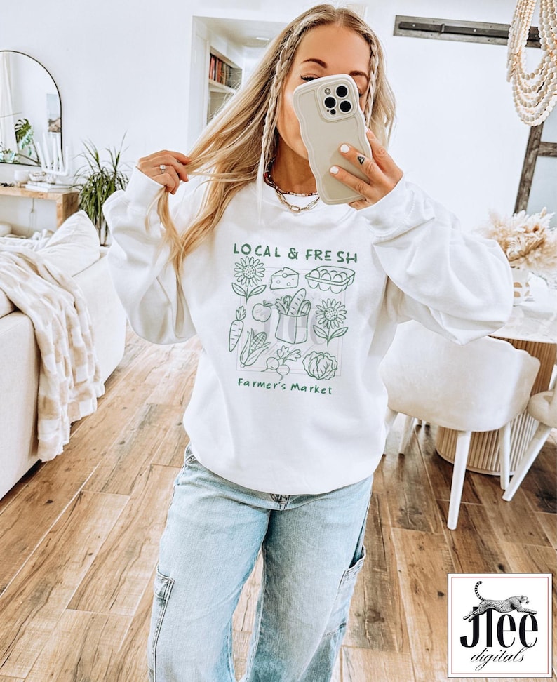 May include: White sweatshirt with the text "LOCAL & FRESH" and a green illustration of farmer's market produce. The design includes sunflowers, cheese, eggs, carrots, apples, a basket of vegetables, and a cabbage. The words "Farmer's Market" are below the illustration.