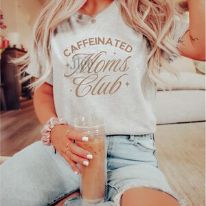 Op de afbeelding: Een vrouw draagt een grijs T-shirt met de tekst "CAFFEINATED Moms Club" in bruine letters. Ze houdt een doorzichtig glas met ijskoffie vast.