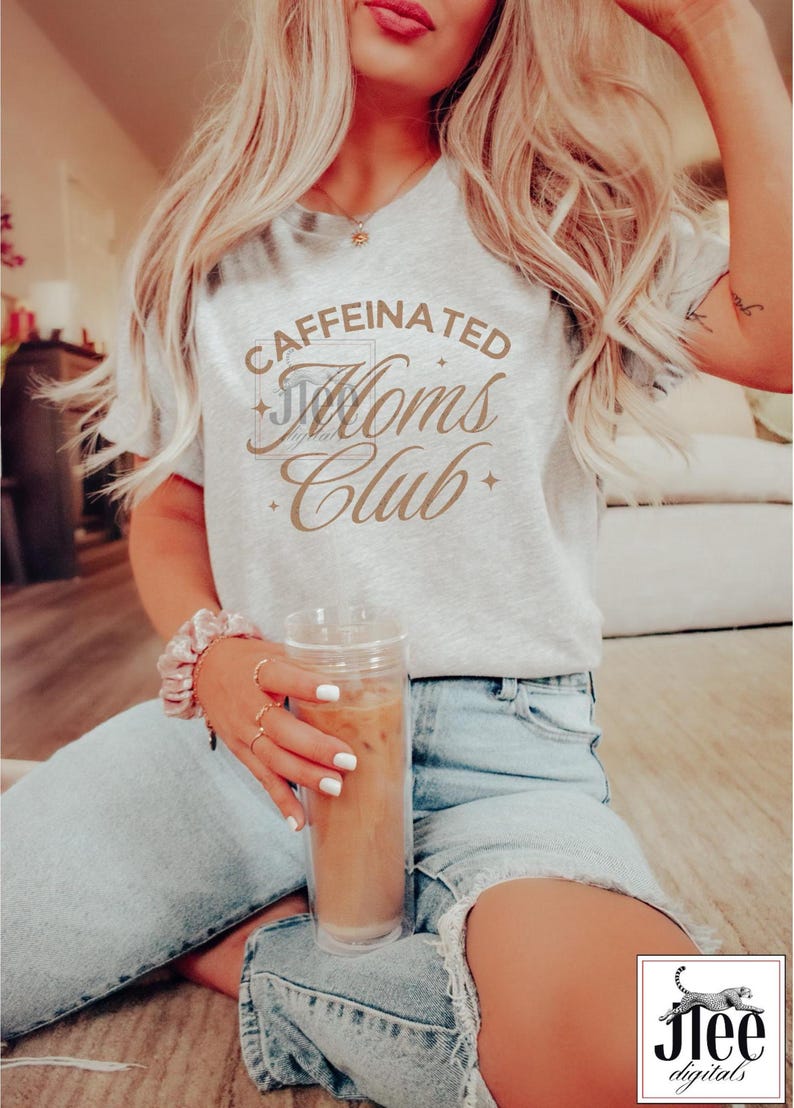 Op de afbeelding: Een vrouw draagt een grijs T-shirt met de tekst "CAFFEINATED Moms Club" in bruine letters. Ze houdt een doorzichtig glas met ijskoffie vast.