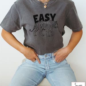 May include: A gray t-shirt with the word "EASY" printed in black letters and a black line drawing of a tiger. The tiger is walking to the right.